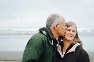 A couple standing on a beach, the man is kissing the woman affectionately on the head.