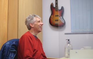 A man sat smiling in a room with a guitar on the wall behind him.